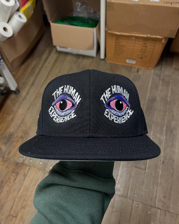 Black cap with embroidered eyes and 'The Human Experience' text held by a person in a warehouse setting.
